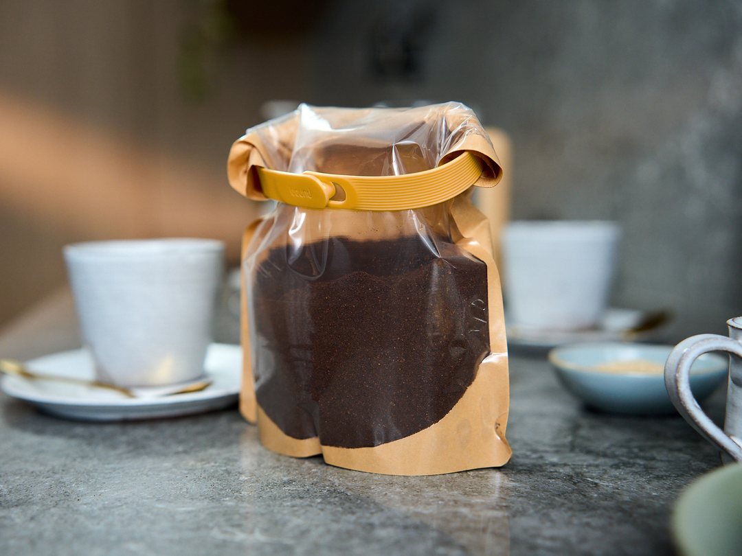 A bag of ground coffee beside two cups, rolled shut with a Mango Looplock